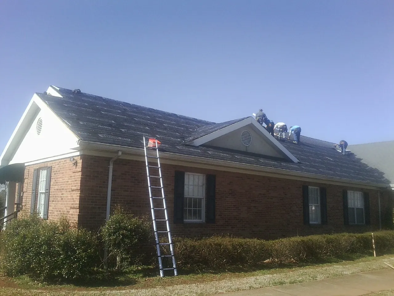 Crew tearing shingles off a brick ranch roof with a ladder set — Emergency Roof Repair work by our crew in Locust Grove