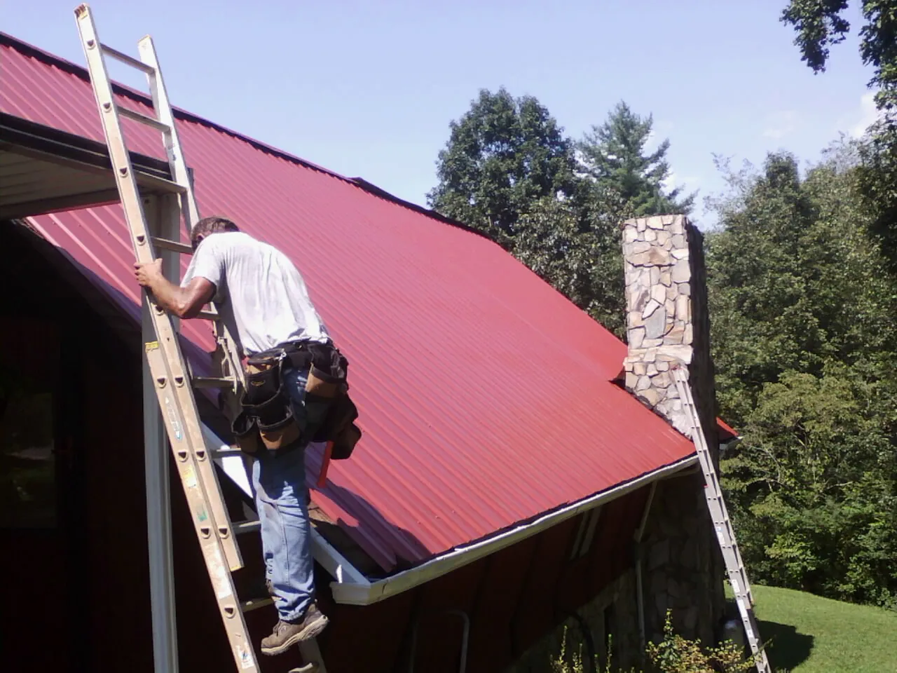 Roofer on a ladder beside a red metal roof and stone chimney — Soffit & Fascia Repair by Roofing Brothers in Locust Grove